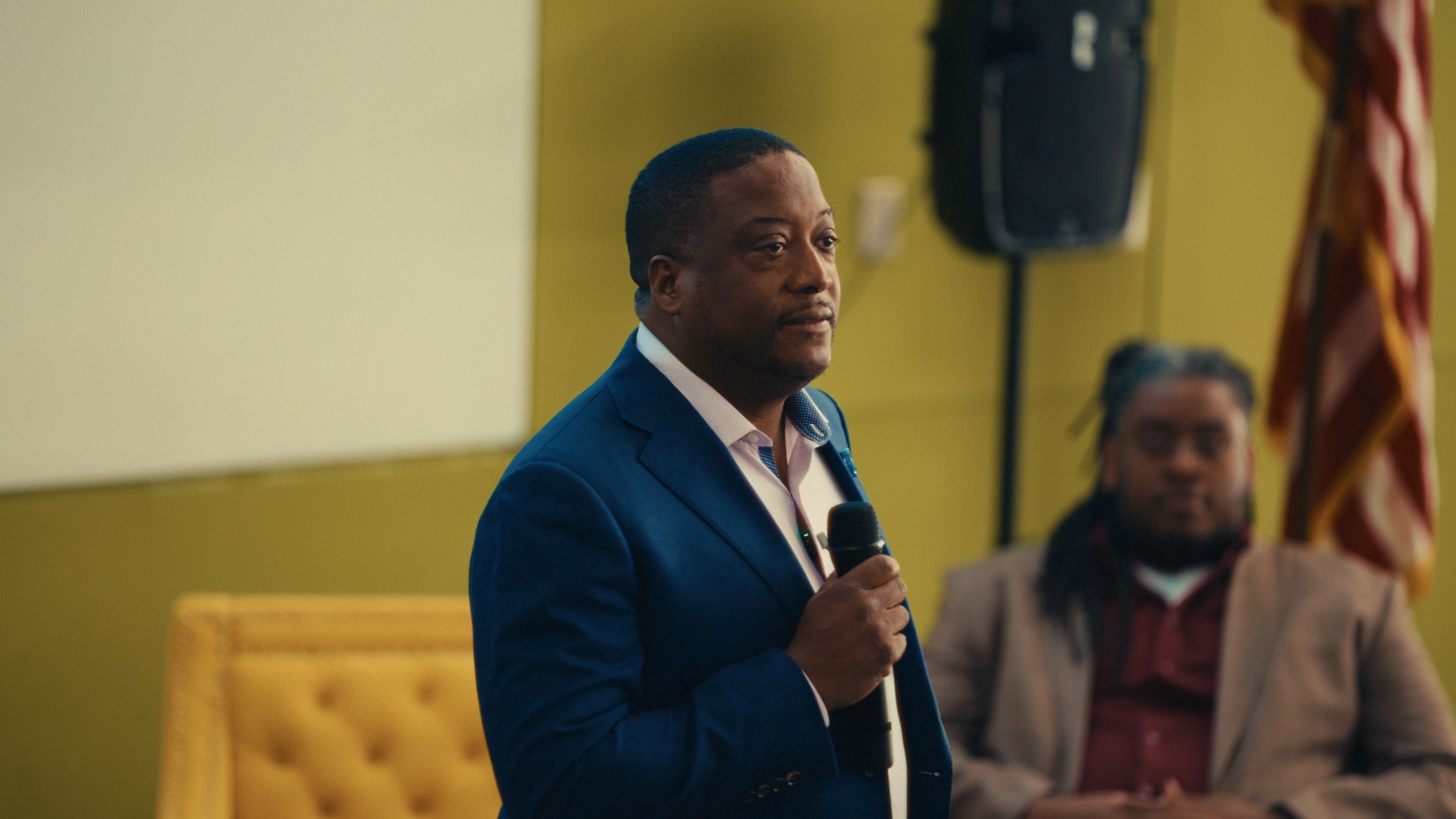 A speaker addresses a room with a microphone during a filmed event